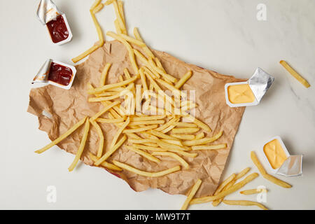 Blick von oben auf die Pommes frites auf zerknittertes Papier mit Behältern von Saucen auf Weiß Stockfoto