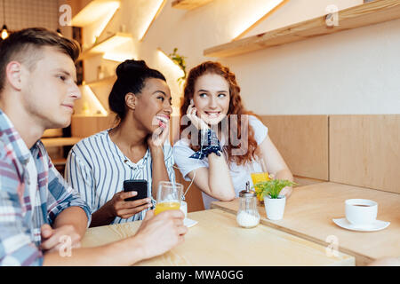 Lächelnden jungen multiethnischen Freunde in Kopfhörer Musik hören mit Smartphone, während im Cafe sitzen Stockfoto