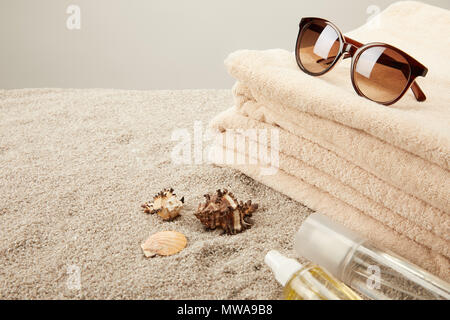 Nahaufnahme Blick auf Stapel von Bettwäsche, Sonnenbrille, das UV-Öl und Muscheln im Sand auf grauem Hintergrund Stockfoto
