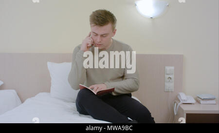Junge casual Geschäftsmann Gespräch per Telefon und schriftlich in Tagebuch konzentriert, Arbeiten mit Notizen im Hotel Zimmer Stockfoto
