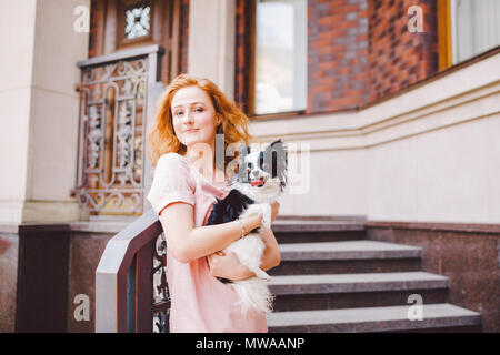 Eine schöne junge Frau mit roten langen Haaren hält ein kleines, süßes Lustig big-eyed Hund von zwei Blumen, ein schwarz-weißes Tier der Rasse der hichuahua Stockfoto