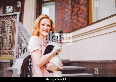 Eine schöne junge Frau mit roten langen Haaren hält ein kleines, süßes Lustig big-eyed Hund von zwei Blumen, ein schwarz-weißes Tier der Rasse der hichuahua Stockfoto