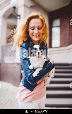 Eine schöne junge Frau mit roten langen Haaren hält ein kleines, süßes Lustig big-eyed Hund von zwei Blumen, ein schwarz-weißes Tier der Rasse der hichuahua Stockfoto