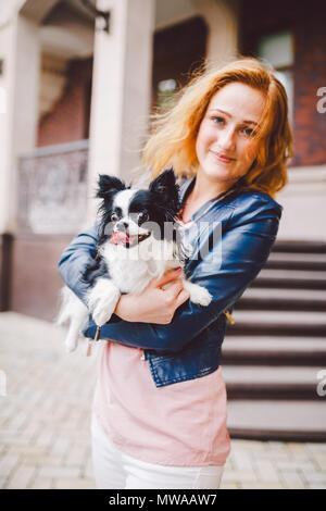 Eine schöne junge Frau mit roten langen Haaren hält ein kleines, süßes Lustig big-eyed Hund von zwei Blumen, ein schwarz-weißes Tier der Rasse der hichuahua Stockfoto