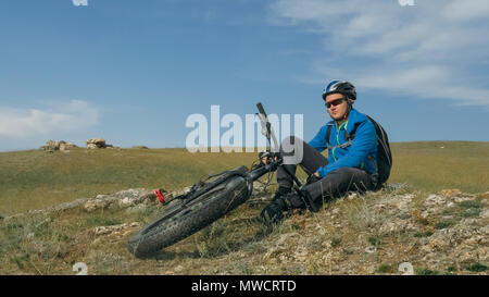 Fat bike auch fatbike oder Fat Tire Bike im Sommer fahren durch die Hügel. Der Kerl sitzt auf einem steinigen Sandstrand und grasbewachsenen Hügel nach einer Reise. Er rest Stockfoto