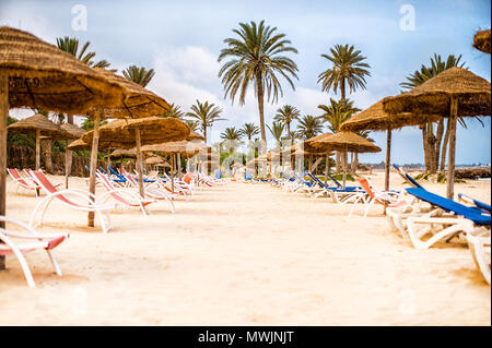 Sonnenliegen und Sonnenschirme Sonnenschirme aus Stroh und Palmen auf Schnee - weißer Sand am Strand in Tunesien Stockfoto