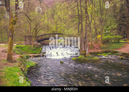 Holzbrücke mit Wasserfall im Naturpark Vrelo Bosne in der Nähe von Sarajevo, Bosnien und Herzegowina. Vintage Style. Stockfoto
