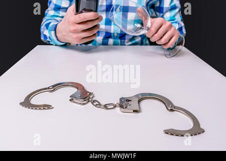 Es gibt Handschellen auf den Tisch, im Hintergrund ein Mann hält eine Flasche Wein und einem Glas. Dem Alkohol verfallen, Bruch mit Alkohol. Das Konzept o Stockfoto