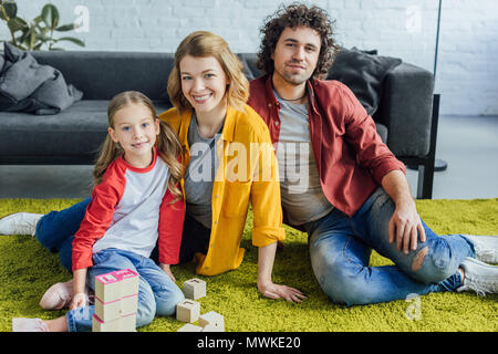 Glückliche Familie sitzt auf dem Teppich und lächelt in die Kamera während des Spielens mit Holzklötzen zu Hause Stockfoto