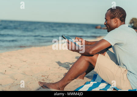 Seitenansicht der lächelnden african american man mit digitalen Tablet beim Sitzen am Sandstrand Stockfoto