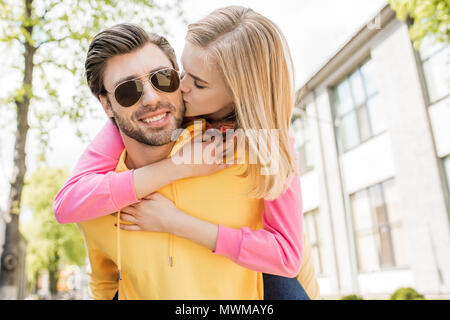 Stilvolle Mann in Sonnenbrille tun piggyback Ride, Freundin, während Sie ihn Küssen in der Backe Stockfoto