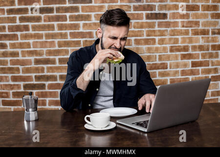 Junger Mann mit Laptop beim Essen Sandwich und Kaffee trinken zum Frühstück bärtigen Stockfoto