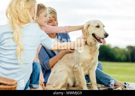 Glückliche junge Eltern mit süßen kleinen Tochter streicheln Hund im Park sitzen Stockfoto