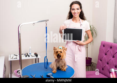 Lächelnd professional groomer Holding leere Zeichen, während in der Nähe von süßen Yorkshire Terrier Hund in Pet Salon stehen Stockfoto