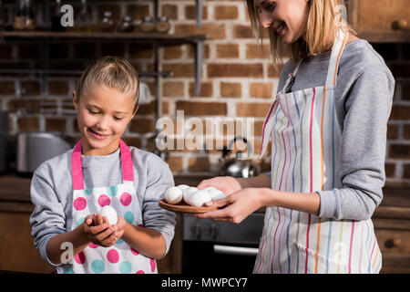 Portrait von kleinen Mädchen und Mutter in Schürzen mit rohem Huhn Eier während zusammen kochen Stockfoto