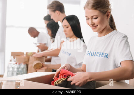 Junge Attraktive freiwillige setzen Kleidung in das Kästchen für Nächstenliebe, während ihre Kollegen im Hintergrund Stockfoto