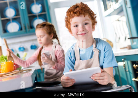 Süße kleine Jungen mit digitalen tablet Lächeln auf die Kamera beim Kind mit Smartphone kochen hinter Stockfoto