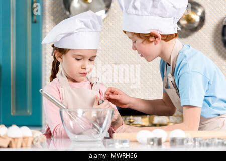 Schöne kleine Kinder in Kochmützen und Schürzen zusammen kochen in der Küche Stockfoto