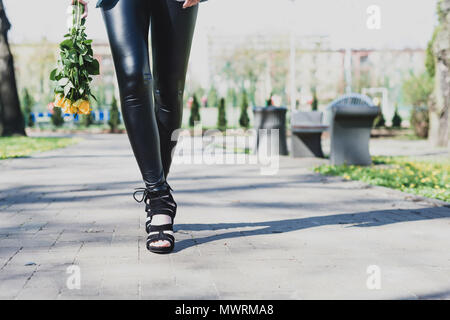 Beine Blick einer Frau mit Blumen. Das Mädchen mit den schwarzen Hosen bewegt sich vor ihr mit gelben Rosen in den Händen. Frauen in Schwarz High Heels re Stockfoto