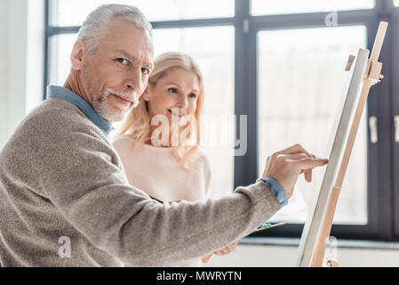 Älterer Mann an der Kamera schaut beim Stehen in der Nähe der lächelnde Frau und Staffelei bei art Klasse Stockfoto