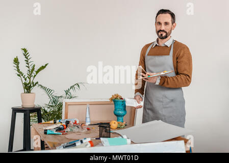 Schöne reife männliche Künstler in Schürze holding Palette und lächelnd an Kamera Stockfoto