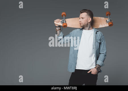 Hübscher junger Mann mit Tattoos holding Skateboard und weg schauen auf Grau isoliert Stockfoto