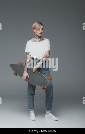 Schöne junge Frau mit Tattoos skateboard halten und Wegschauen auf Grau Stockfoto