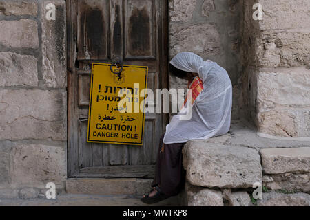 Ein äthiopisch-orthodoxer Christ Pilger sitzt vor dem geschlossenen Eingang zur St. Michael Kirche der äthiopisch orthodoxen Kammer am Parvis oder Parvise der Kirche des Heiligen Grabes in der christlichen Viertel Altstadt Ost-Jerusalem Israel. Die Kirche wurde geschlossen, nachdem ihr Dach durch den Bau auf dem Dach Kloster von der griechischen Kirche eingestürzt war, Streit zwischen den Kirchen in den Weg der israelischen Regierung Bemühungen, mit dem Wiederaufbau voranzukommen. Stockfoto