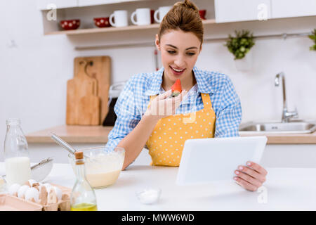 Attraktive junge Frau essen Erdbeeren und mit Tablet in der Küche beim Kochen Stockfoto