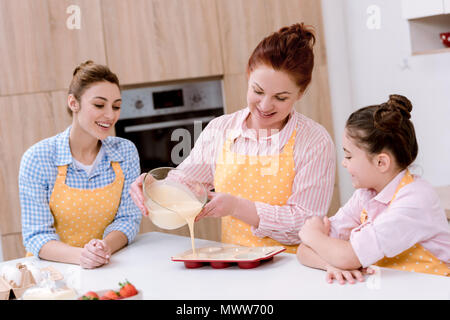 Drei Generationen von Frauen Gießen Teig in Backformen für Cupcakes in Küche Stockfoto