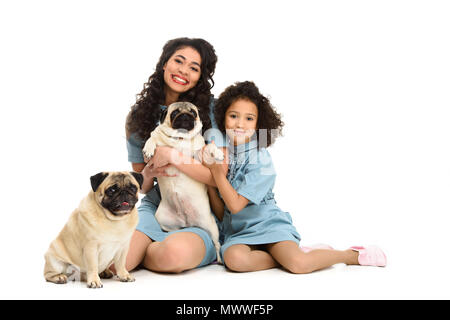 Glückliche junge Mutter und Tochter sitzen auf dem Boden mit zwei adorable Möpse isoliert auf weißem Stockfoto