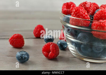 Nahaufnahme, Glas Schüssel voll von Blaubeeren mit Himbeeren auf, einige von ihnen auf grau Holz Schreibtisch verschüttet. Stockfoto
