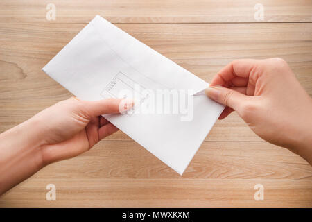 Woman's Hand hält und öffnet einen weißen Umschlag oder Brief Briefumschläge auf einem hölzernen Schreibtisch Draufsicht Hintergrund. Stockfoto
