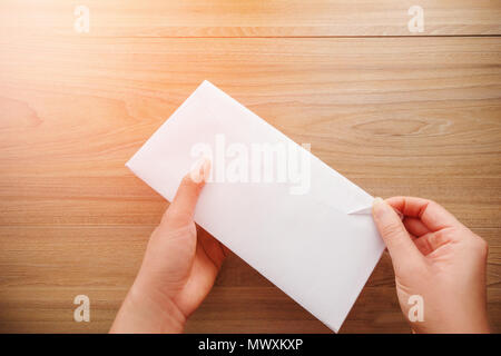 Woman's Hand hält und öffnet einen weißen Umschlag oder Brief Briefumschläge auf einem hölzernen Schreibtisch mit natürlichem Sonnenlicht am Morgen, Ansicht von oben Hintergrund. Stockfoto