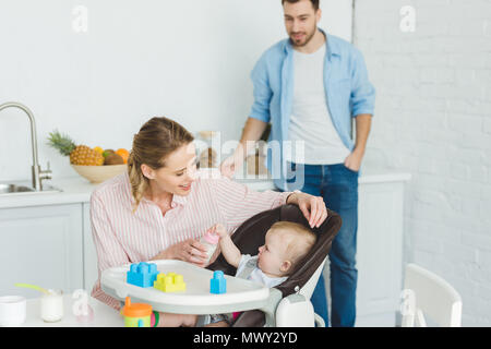 Mutter geben Sie die Flasche auf den neugeborenen Tochter in Baby Stuhl während Vater hinter Stockfoto