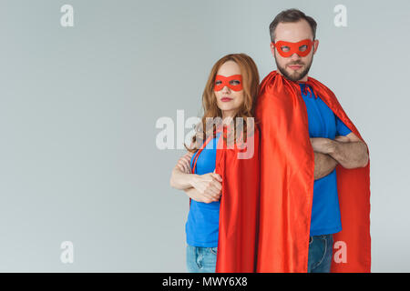 Super Paar in Masken und Kleider stehen mit verschränkten Armen und Kamera isoliert auf Grau Stockfoto