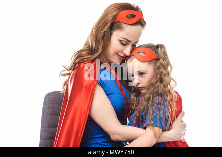 Lächelnde Mutter in Superhelden Kostüm umarmen kleine Tochter in Maske und Umhang isoliert auf weißem Stockfoto