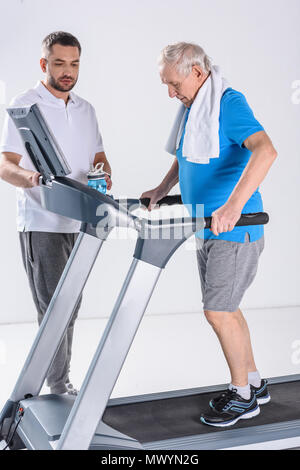 Rehabilitation Therapeuten Unterstützung älterer Mann mit Handtuch Ausübung auf Laufband isoliert auf Grau Stockfoto