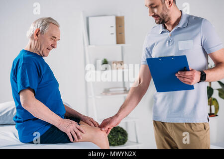 Seitenansicht der Rehabilitation Therapeuten mit Notepad prüfen Senior mans Knie auf Massage Tisch Stockfoto