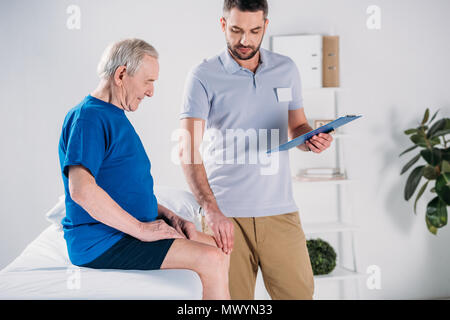 Rehabilitation Therapeuten mit Notepad prüfen Senior mans Knie auf Massage Tisch Stockfoto