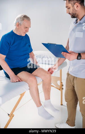 Rehabilitation Therapeuten mit Notepad prüfen Senior mans Knie auf Massageliege auf grauem Hintergrund Stockfoto