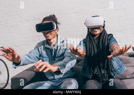 Junge lächelnde Freunde sitzen auf der Couch und mit Virtual reality Headsets Stockfoto
