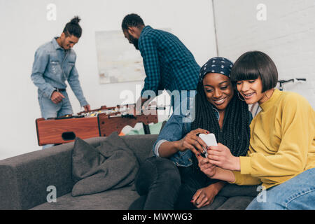 Zwei junge weibliche Freunde sitzen mit Smartphone und zwei junge Männer Spielen Table Soccer hinter Stockfoto