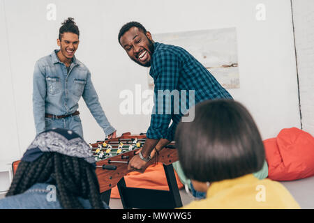 Lachend multiethnischen Freunde Spielen Table Soccer und zwei weibliche Freunde neben Sitzen Stockfoto