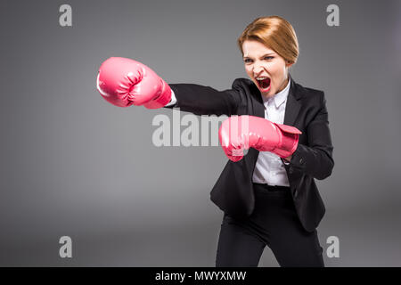 Aggressive Geschäftsfrau kreischend in rosa Boxhandschuhe, isoliert auf Grau Stockfoto