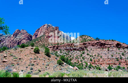 Orange Schmutz Hang mit Red Rock Berge hinter mit grünen Bäumen und Laub unter einem strahlend blauen Himmel. Stockfoto