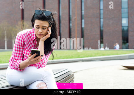Eine schöne junge Frau auf einer Bank sitzen. Hübsches Mädchen in Sonnenbrille sieht am Telefon besorgt. Eine Frau überprüft Ihr Kontostand nach shoppin Stockfoto