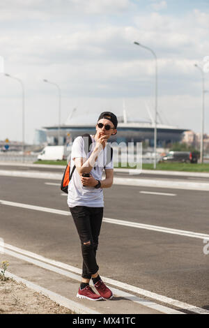 Stilvolle mann Tourist auf der Straße auf den Hintergrund des unscharfen Fußballstadion. Er trug, schwarze Jeans, Schwarzes T-Shirt, schwarze Kappe mit riund Sonnenbrille. Konzept der Fußball, Fans, Fußball-WM. Stockfoto