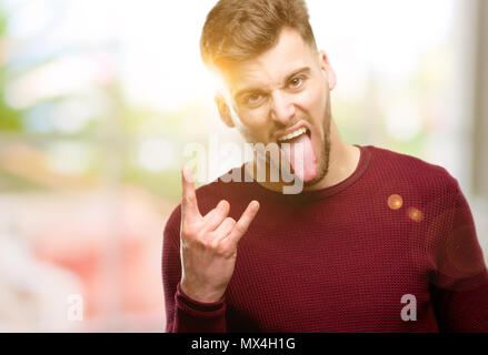 Hübscher junger Mann, Rock Symbol mit Händen, Schreien und feiern Mit herausgestreckter Zunge Stockfoto