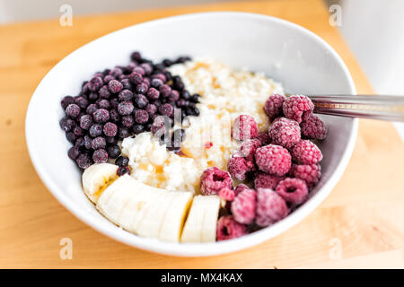 Makro Nahaufnahme von haferflocken Hafer Reis Pudding porridge Bowl mit gefrorenen Beeren, Himbeeren, Heidelbeeren, Bananen und Ahorn Sirup Karamell auf Holz Stockfoto